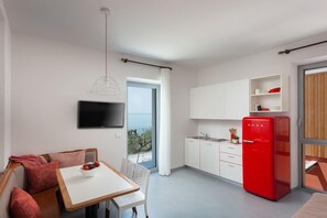 A modern kitchen with a red refrigerator, a dining area with a table and chairs, a TV mounted on the wall, and a view of the sea through a sliding door. - Appartamenti Arca and Ca Mure (Bardolino)