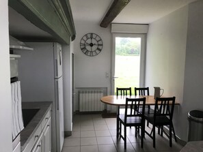 A kitchen with a dining table and chairs, a clock on the wall, and a view of greenery outside. - Single-storey house in the heart of nature (Loison-sur-Créquoise)