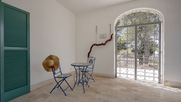 A room with a green shutter door, a table with two chairs, a decorative branch, and a large arched window with a metal gate. - GuestHost - Large room ideal for two, featuring a private bathroom and located in a villa in Borgo Lucia, a complex with a large shared garden. Surrounded by greenery, the accommodation is located in Selva di Fasano, a quiet hillside hamlet in the munici (Selva di Fasano)