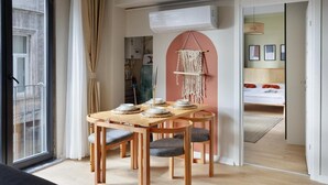 Un soggiorno moderno con un tavolo da pranzo in legno apparecchiato per quattro persone, una grande finestra con tende e vista su una camera da letto con un letto e una lampada a sospensione.
