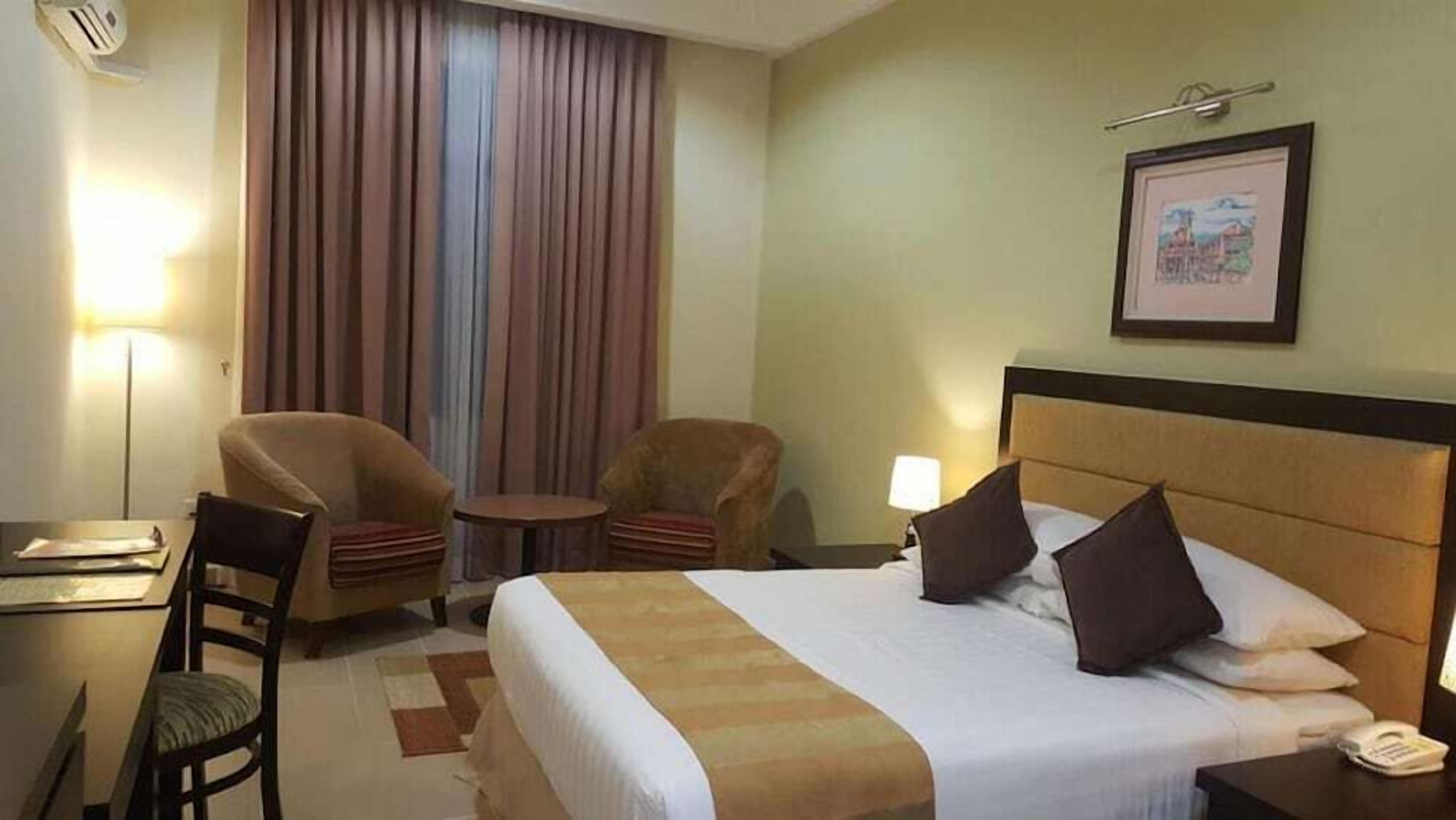 A hotel room with a bed, two armchairs, a desk, a lamp, and a framed picture on the wall.