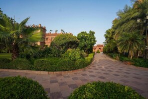 A paved pathway leading to a building with a red facade, surrounded by lush greenery and palm trees. - Marugarh (Jodhpur)