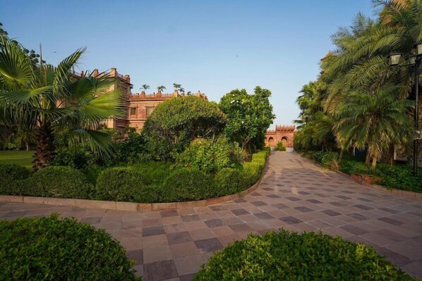 A paved pathway leading to a building with a red facade, surrounded by lush greenery and palm trees. - Marugarh (Jodhpur)
