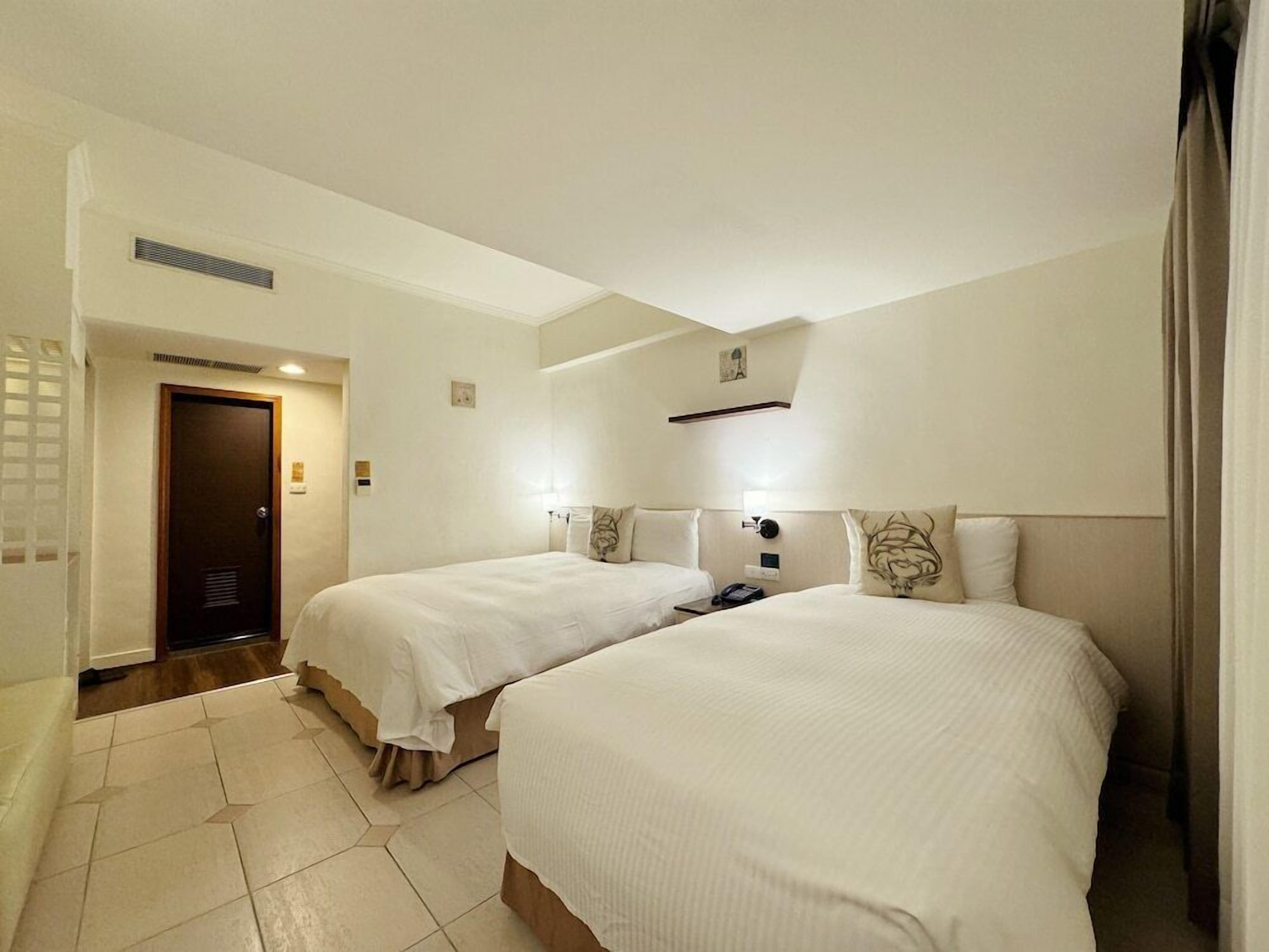 Habitación de hotel con dos camas, una puerta, una lámpara y un pequeño adorno en la pared.