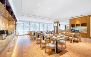 A hotel dining area with multiple tables and chairs, a kitchen area, and a large window providing natural light. - Vienna Hotel (Dongguan)