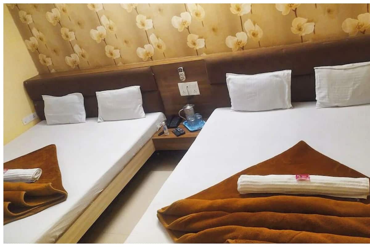 A hotel room with two beds, each with white linens and pillows. There is a wooden nightstand between the beds with a lamp, a phone, and a small table with a glass and a bottle. The headboard has a floral pattern.