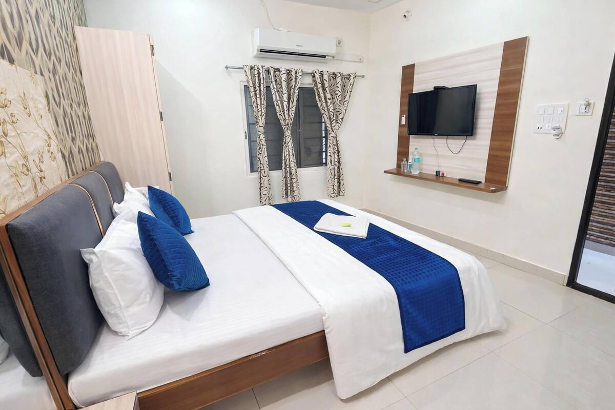A neatly made bed with white and blue bedding, a wooden headboard, and a blue accent pillow. There is a television mounted on the wall, a window with patterned curtains, and a small shelf with a bottle and remote.
