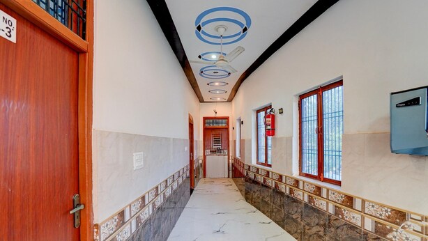 A hallway with a marble floor, a wooden door, a ceiling fan, and a window with a metal grill. - Classic Hotel Siya Raman (Faizabad)