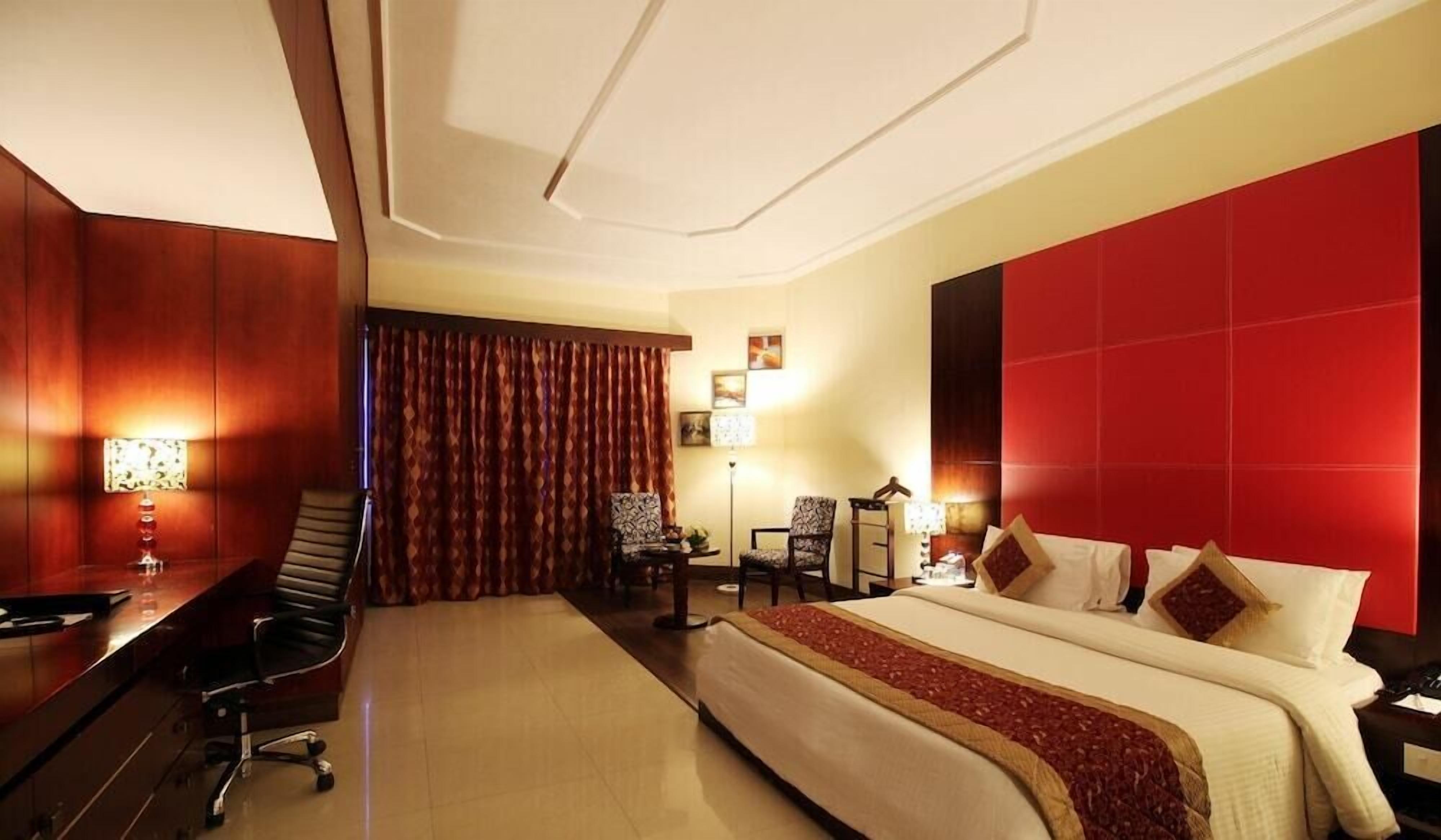 A hotel room with a large bed, a desk, two chairs, a red accent wall, and a ceiling with recessed lighting.