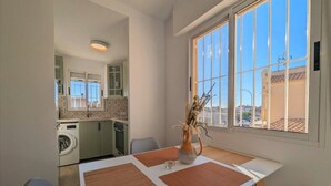Zona pranzo con tavolo, sedie, un vaso con piante essiccate e vista su una cucina ed edifici esterni.