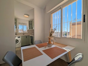 A dining area with a table, chairs, a vase with dried plants, and a view of a kitchen and outside buildings.