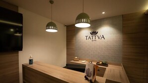 Un área de recepción con un mostrador de madera, dos lámparas colgantes, un televisor montado en la pared y un letrero que dice "TATTVA SPA".
