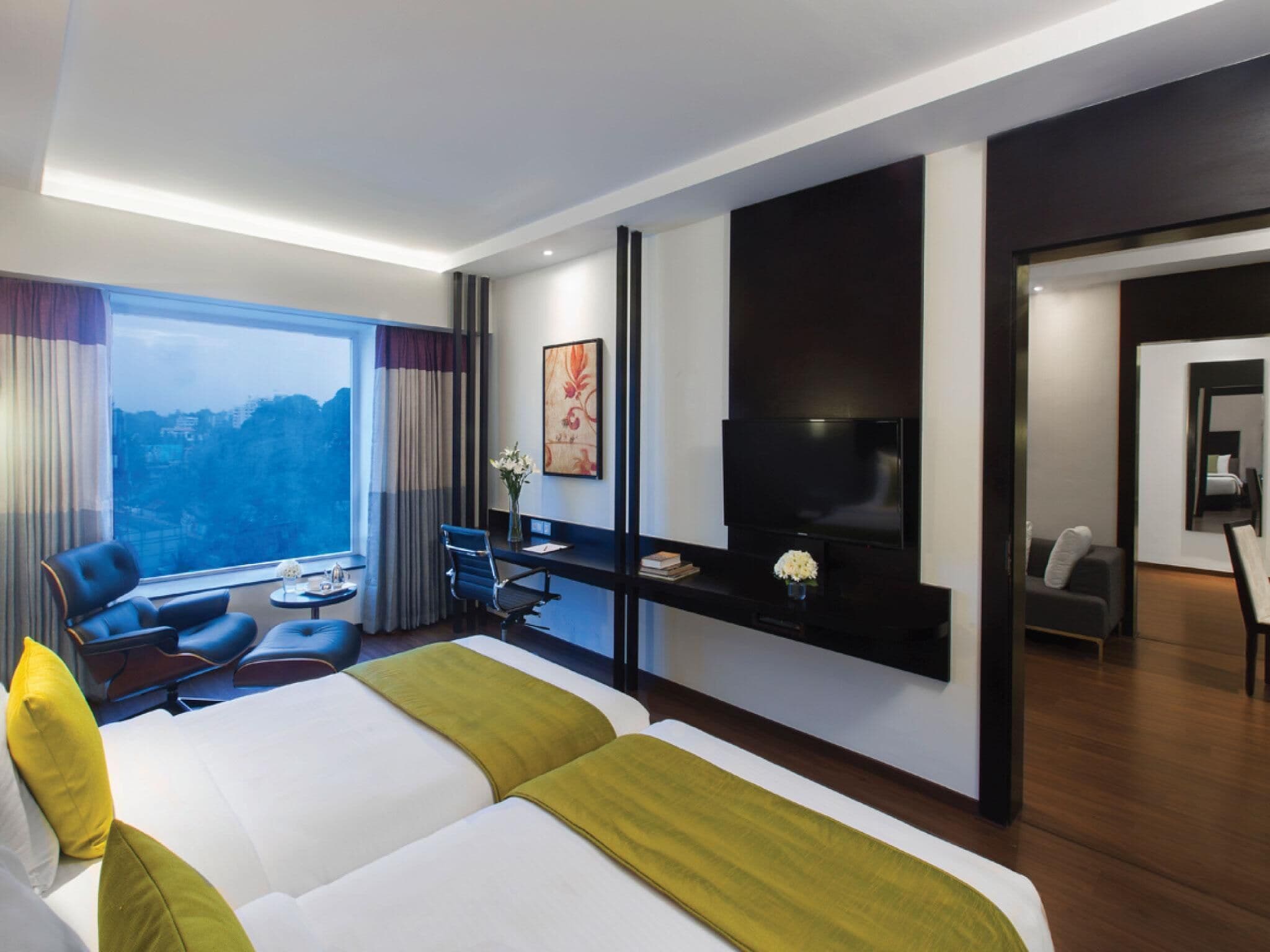 A modern hotel room with a large bed, a desk, a chair, a TV, and a view of the city.