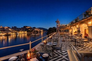 A waterfront restaurant with tables set for dining, overlooking a river with city buildings in the background. - Hotel Sarovar (Udaipur)