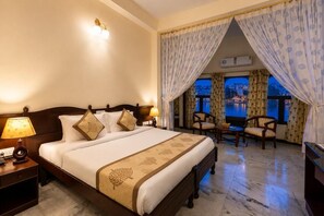 A hotel room with a large bed, two bedside lamps, a desk with a lamp, a chair, and a view of a cityscape through a window with curtains. - Hotel Sarovar (Udaipur)