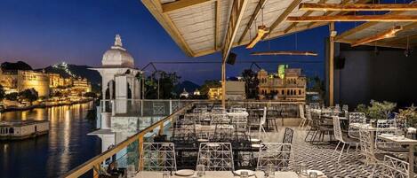 An outdoor dining area with tables and chairs, overlooking a river and historic buildings.