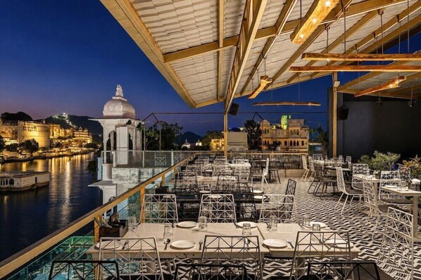 An outdoor dining area with tables and chairs, overlooking a river and historic buildings. - Hotel Sarovar (Udaipur)