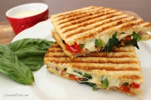 A grilled sandwich with visible ingredients like spinach, tomatoes, and cheese, served on a white plate with a red cup of sauce in the background. - Hotel Sarovar (Udaipur)