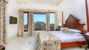 A hotel room with a bed, a nightstand, a lamp, and a view of mountains through large windows. - Fateh Garh (Udaipur)