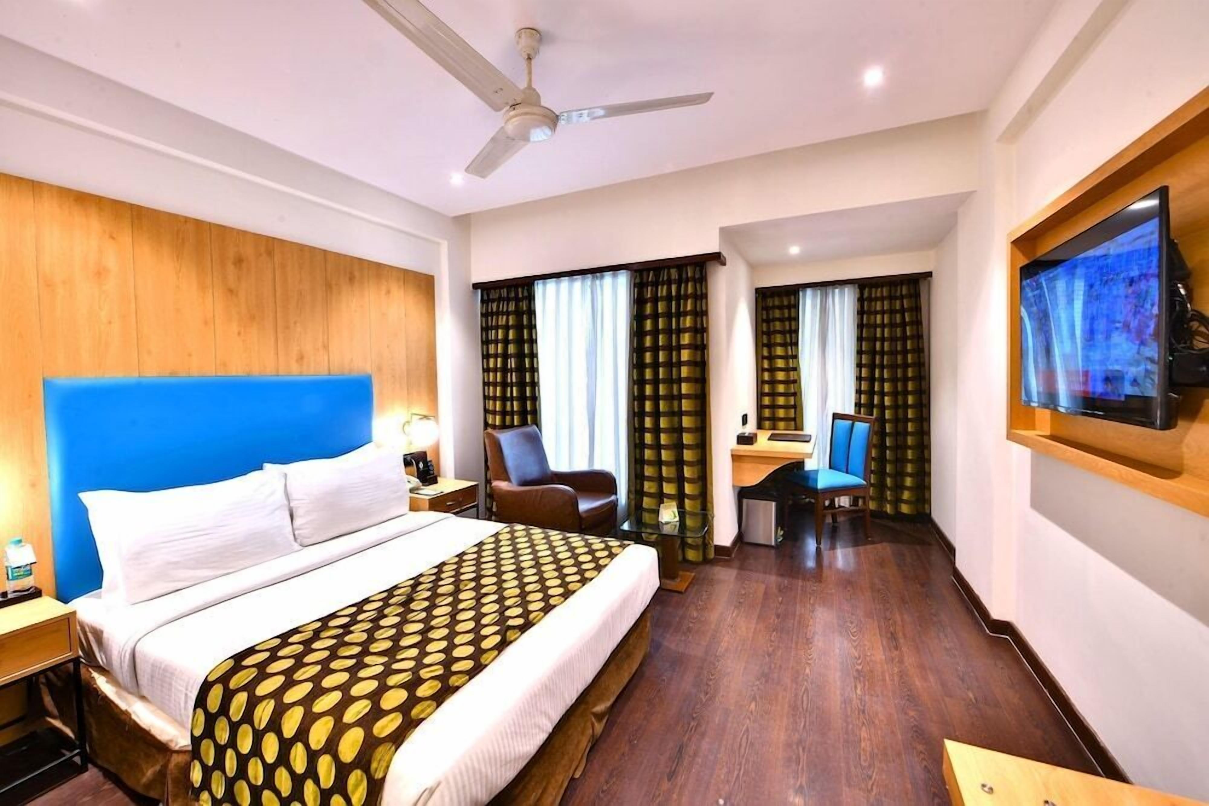 A hotel room with a large bed, a desk, a chair, a television, and a ceiling fan.