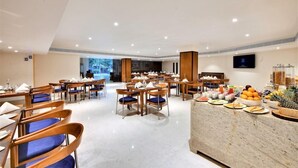 Ein geräumiger Hotel-Speiseraum mit mehreren Tischen und Stühlen, einer Speiseauslage mit verschiedenen Speisen und einem großen Bildschirm, der eine schöne Landschaft zeigt.