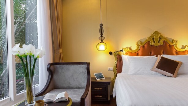Une chambre d’hôtel avec un lit, une chaise, une table de chevet avec un livre et une tasse, une petite table de nuit avec une lampe, et une fenêtre avec des rideaux.