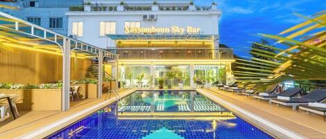 A modern hotel with a rooftop pool, outdoor seating, and a well-lit entrance.