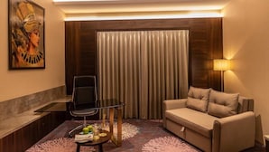 Una habitación de hotel moderna con sofá, una mesa de vidrio con manzanas, una silla, un cuadro en la pared y un ventanal con cortinas.
