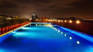 A nighttime swimming pool with a wooden deck and a cityscape in the background. - E Square the Fern Pune (Pune)