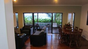 A spacious living area with a wooden floor, a sofa, a dining table with chairs, and a view of greenery through large glass doors. - Raina Beach Apartments (Rarotonga)