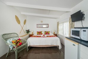 Un dormitorio con cama, una silla de mimbre verde, pared blanca, televisor de pantalla plana, microondas y ventana con cortinas.