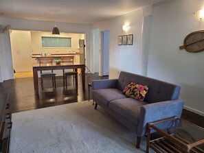 Una sala de estar con un sofá gris, una mesa de comedor con sillas, una cocina al fondo y un ventanal grande.