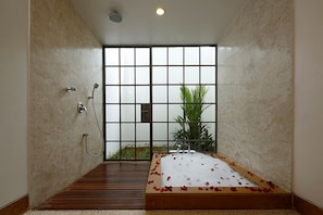 A spa room with a wooden floor, a bathtub with rose petals, a shower, and a large window with a view of greenery.