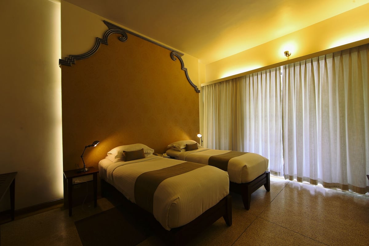 Habitación de hotel con dos camas, una mesita de noche con lámpara, un ventanal con cortinas y un zócalo decorativo.