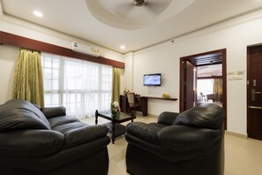A hotel room with a sofa, armchairs, a TV, a desk, and a window with curtains. - NO 18 Hotel (Kochi)