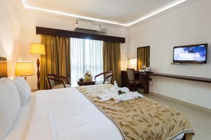 A hotel room with a bed, two chairs, a desk, a television, and an air conditioning unit.