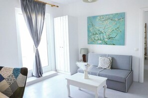 A room with a grey sofa, a white coffee table, a painting of a tree, and a window with curtains. - La Stella Hotel (Mykonos)