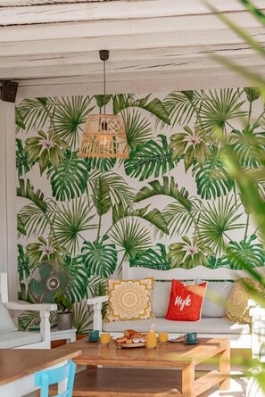 A room with a tropical leaf wallpaper, a wicker hanging light, a sofa with patterned cushions, and a dining table with breakfast items. - La Stella Hotel (Mykonos)