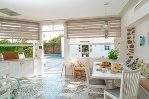 Un luminoso comedor con una mesa dispuesta para dos personas, un sofá y vistas a una piscina y vegetación exterior.