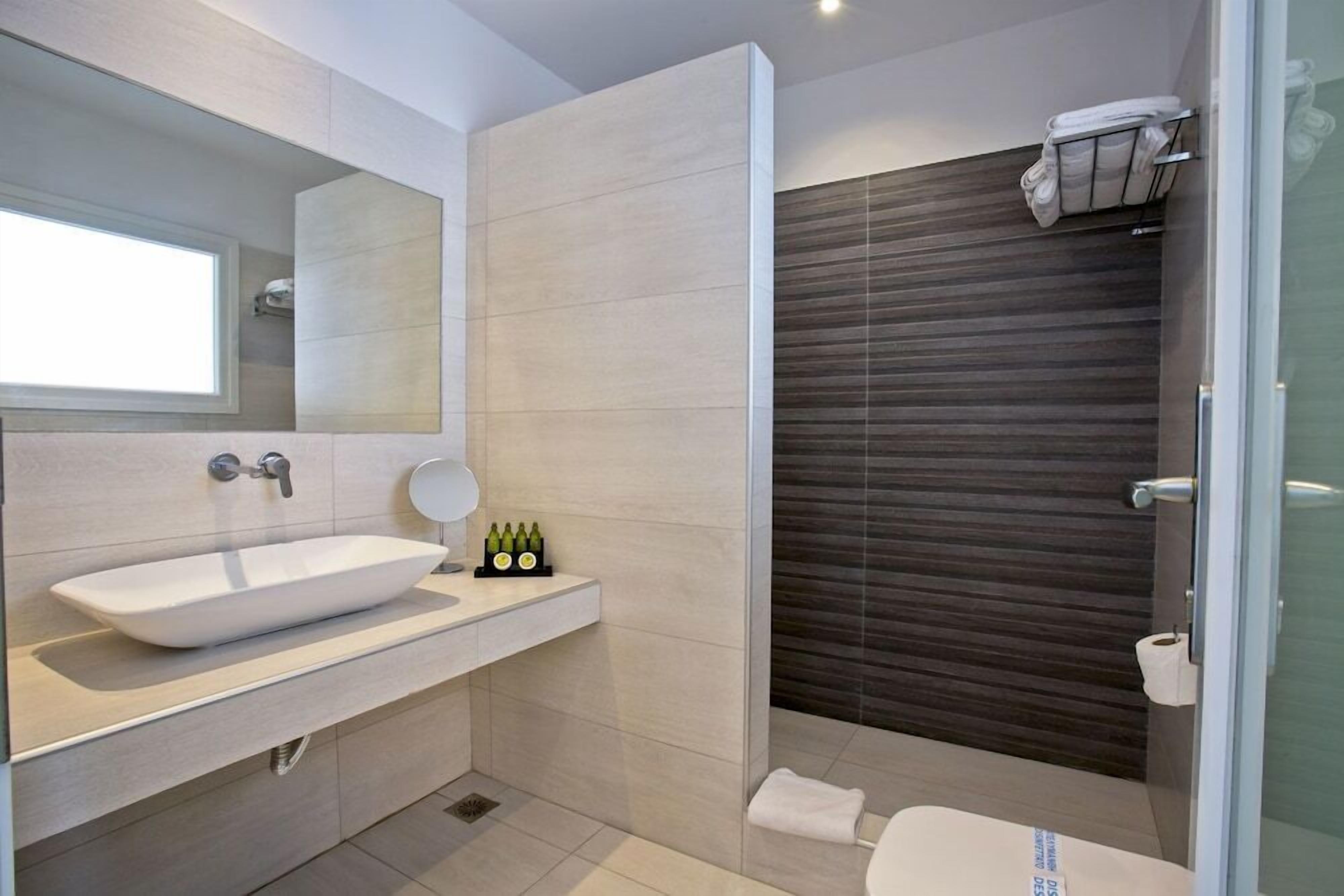 A modern bathroom with a large vessel sink, a mirror, and a shower area with a dark wood panel wall.