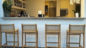A bar area with three wicker chairs, a shelf with bottles, and a potted plant. - Anna Maria Mykonos Hotel (Mykonos)