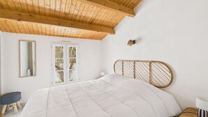 Ett sovrum med ett träpanelat tak, en säng med en vit sänggavel, ett sängbord med en lampa, en spegel och ett fönster med utsikt över träd.