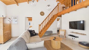 Ett rymligt vardagsrum med ett högt träpanelat tak, en trätrappa, en stor platt-TV som är monterad på väggen och en mysig sittgrupp med en soffa och ett soffbord.