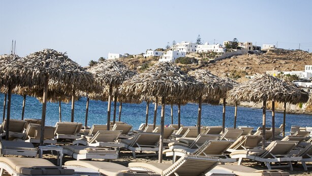 A beach with rows of sun loungers and thatched umbrellas, a clear blue sea, and white buildings on a hillside in the background. - Asteri Hotel Ornos (Mykonos)