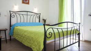 A bed with a green and blue quilt, a small bedside table, a wall-mounted light, a framed picture, and a green curtain. - Asteri Hotel Ornos (Mykonos)