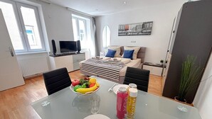 A modern living room with a sofa, a dining table with fruit and drinks, and a large window.