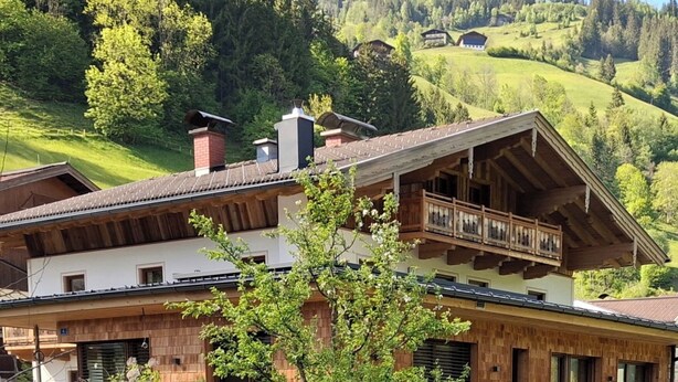 Un chalet de dos plantas con fachada de madera, un balcón en el piso superior y vistas a una ladera verde al fondo.