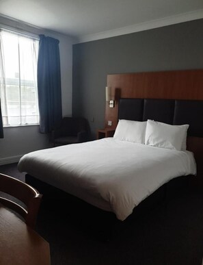 Ein Hotelzimmer mit einem großen Bett, einem Holzkopfteil, einem Nachttisch, einem Sessel und einem Fenster mit Vorhängen.