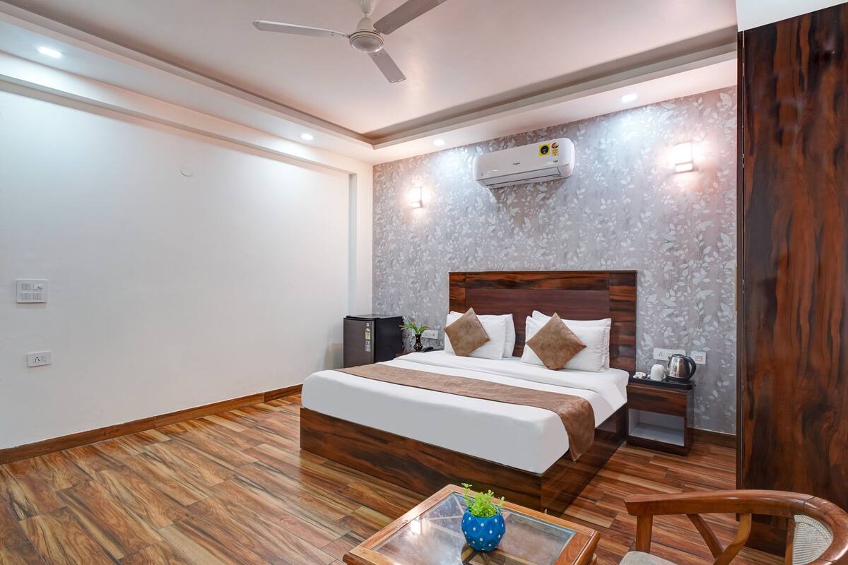 Habitación de hotel con piso de madera, cama con ropa blanca y almohadas marrones, mesita de noche con lámpara y ventilador de techo.