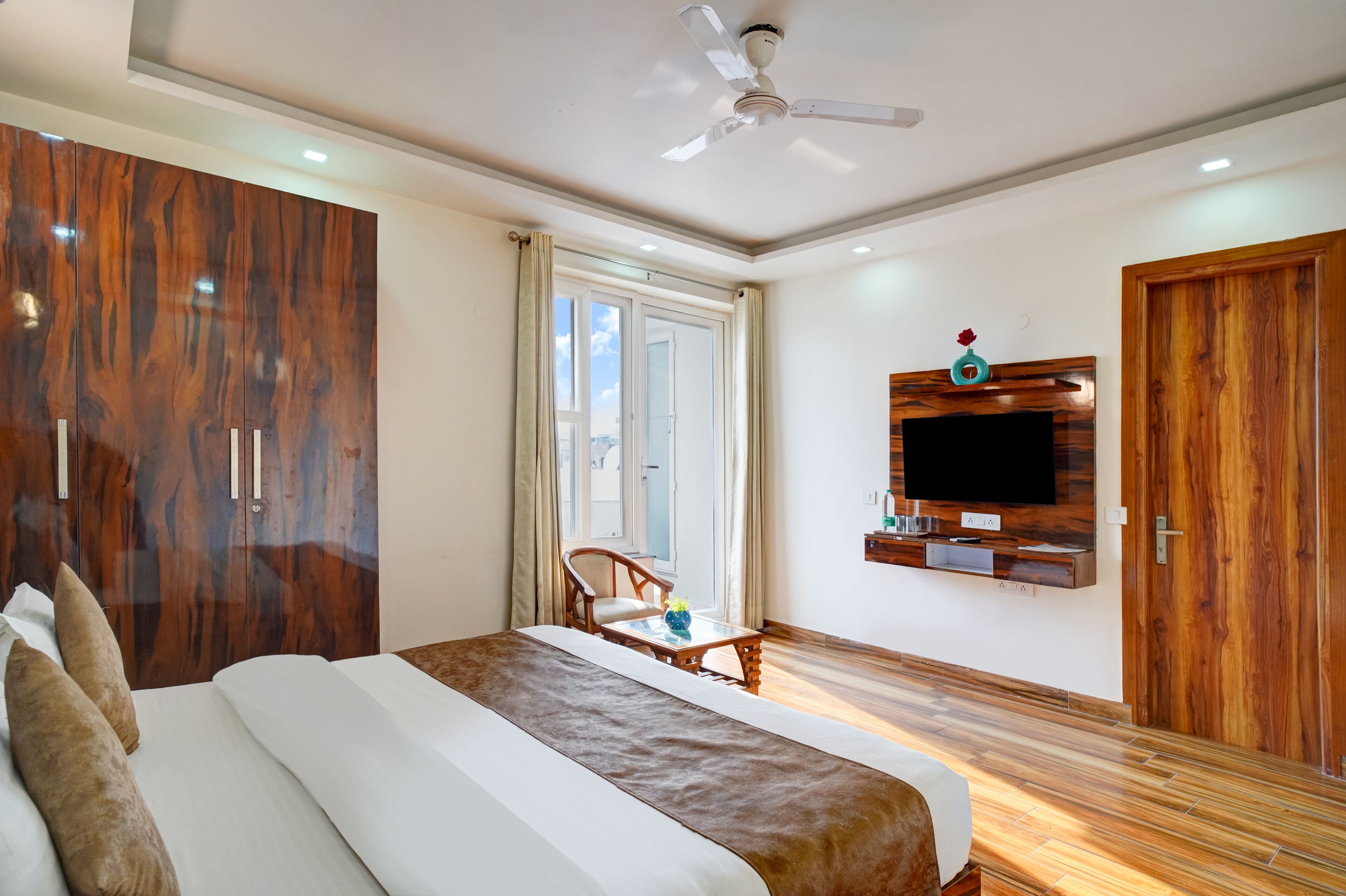 A bedroom with a bed, a wooden wardrobe, a TV mounted on the wall, and a ceiling fan.
